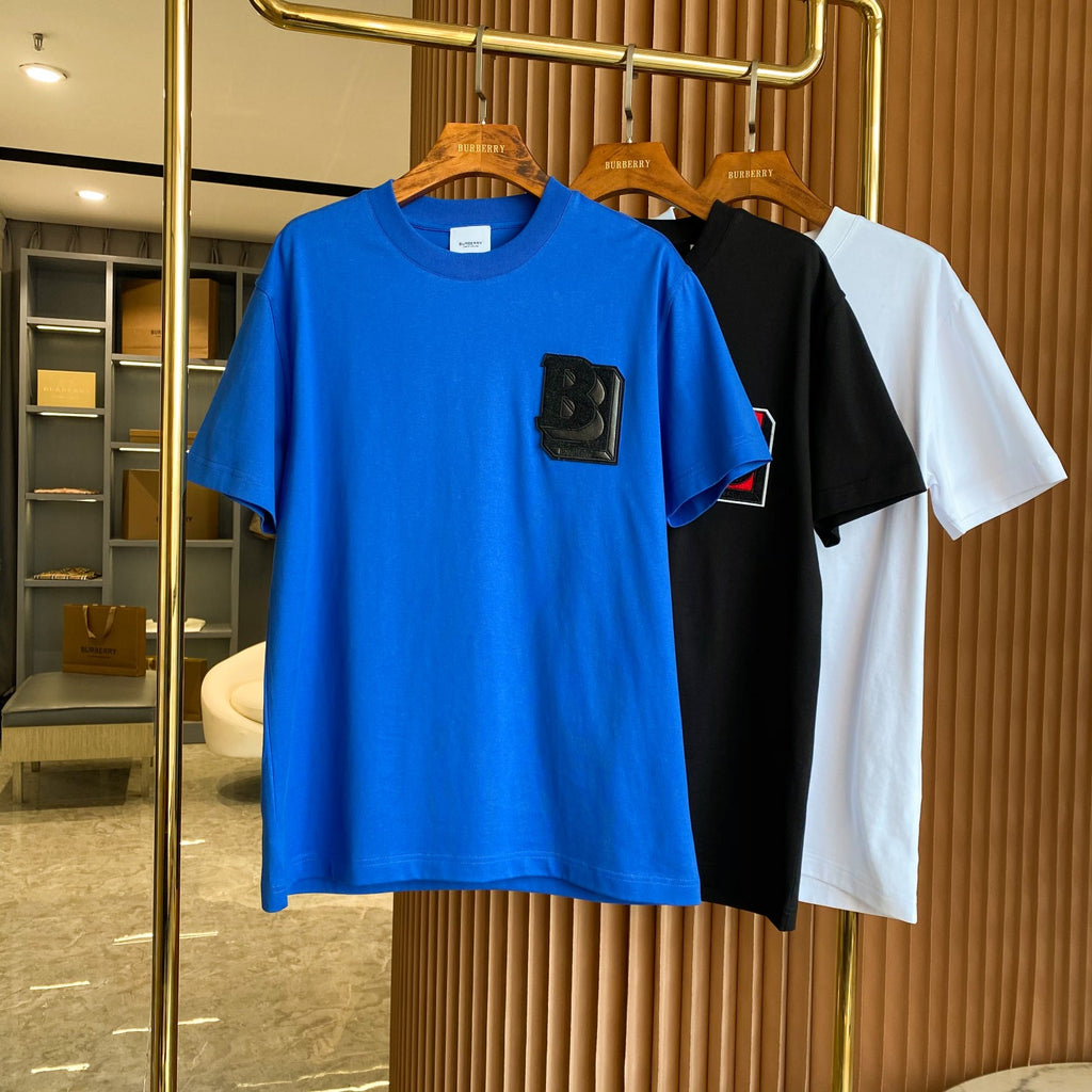 Blue short-sleeve T-shirt with black B logo patch on rack with other colors.