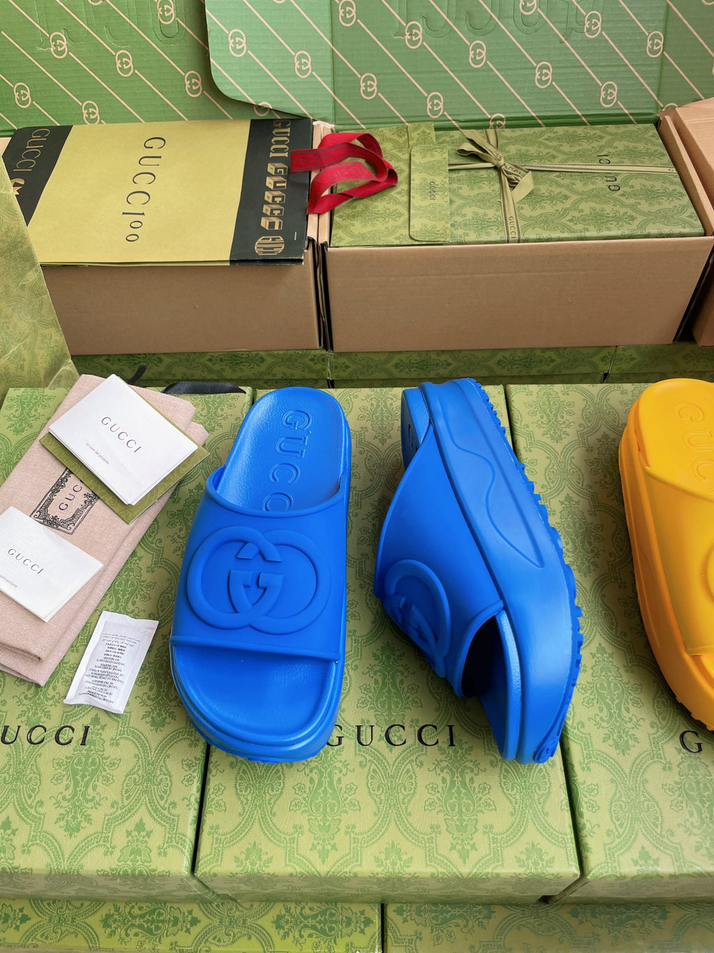Gucci slide sandals in royal blue with embossed logo footbed; yellow pair partly visible.
