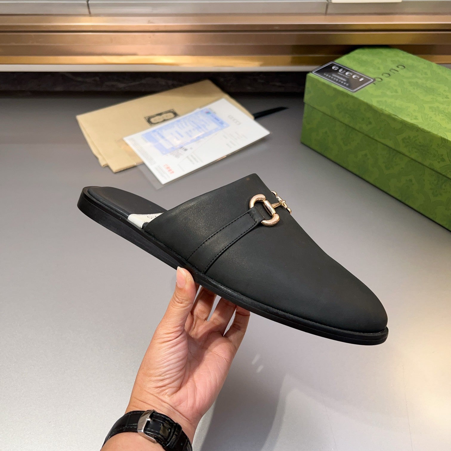 Gucci black leather mule sandal held in hand, gold-tone buckle detail on upper