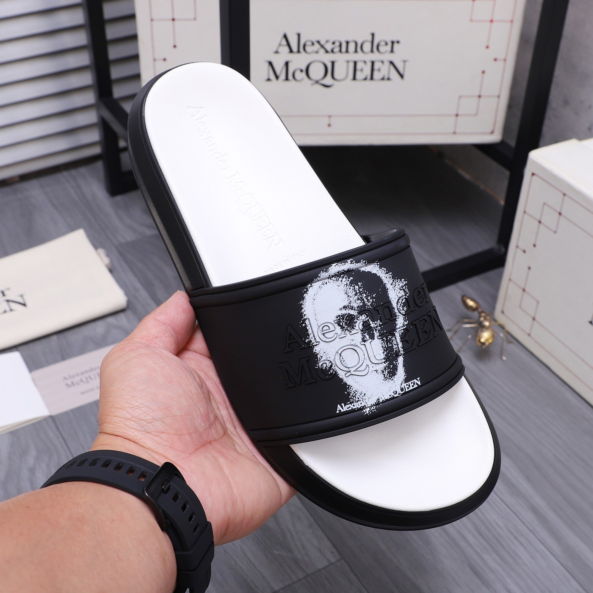 Black slide sandal held in hand; strap has white portrait graphic on black band.