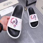 Alexander McQueen slide sandals held in hand; white uppers with red/blue skull print