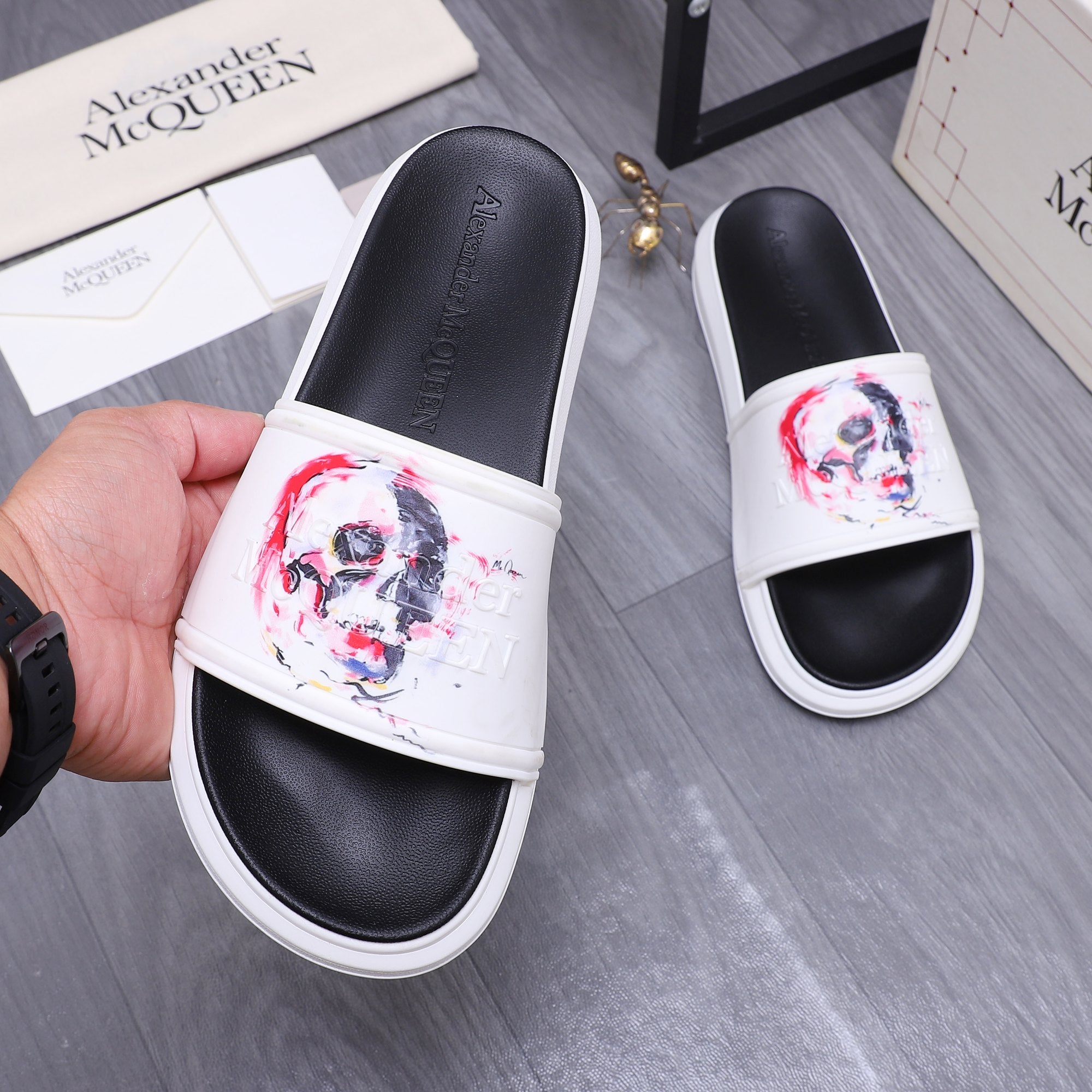 Alexander McQueen slide sandals held in hand; white uppers with red/blue skull print