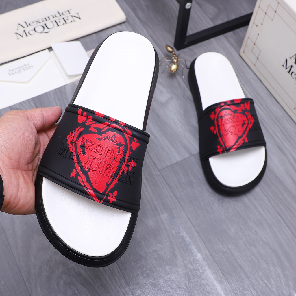 Black slide sandal with red heart-like graphic strap and white footbed, held in hand
