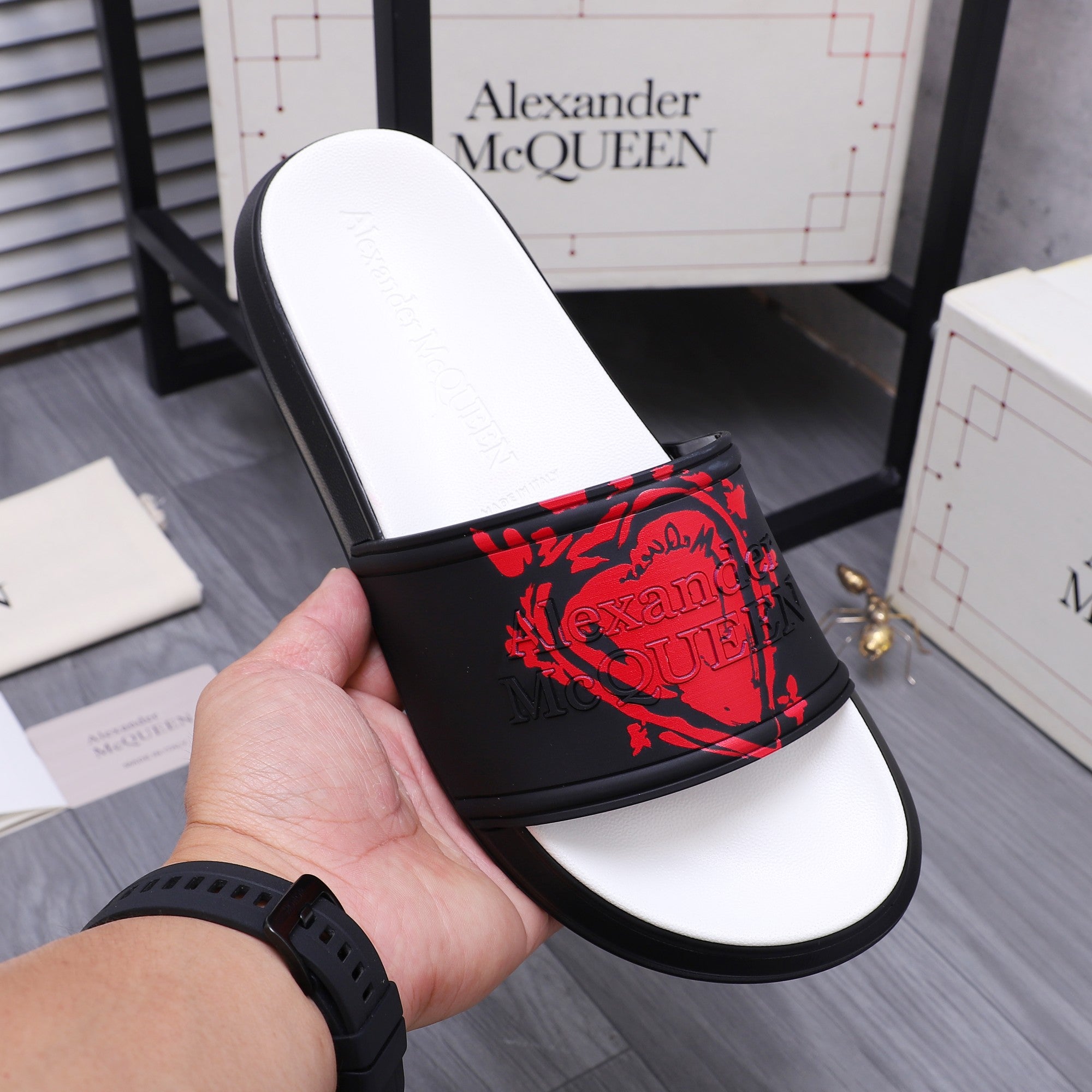 Black slide sandal with red graphic strap and white footbed, hand holding the shoe