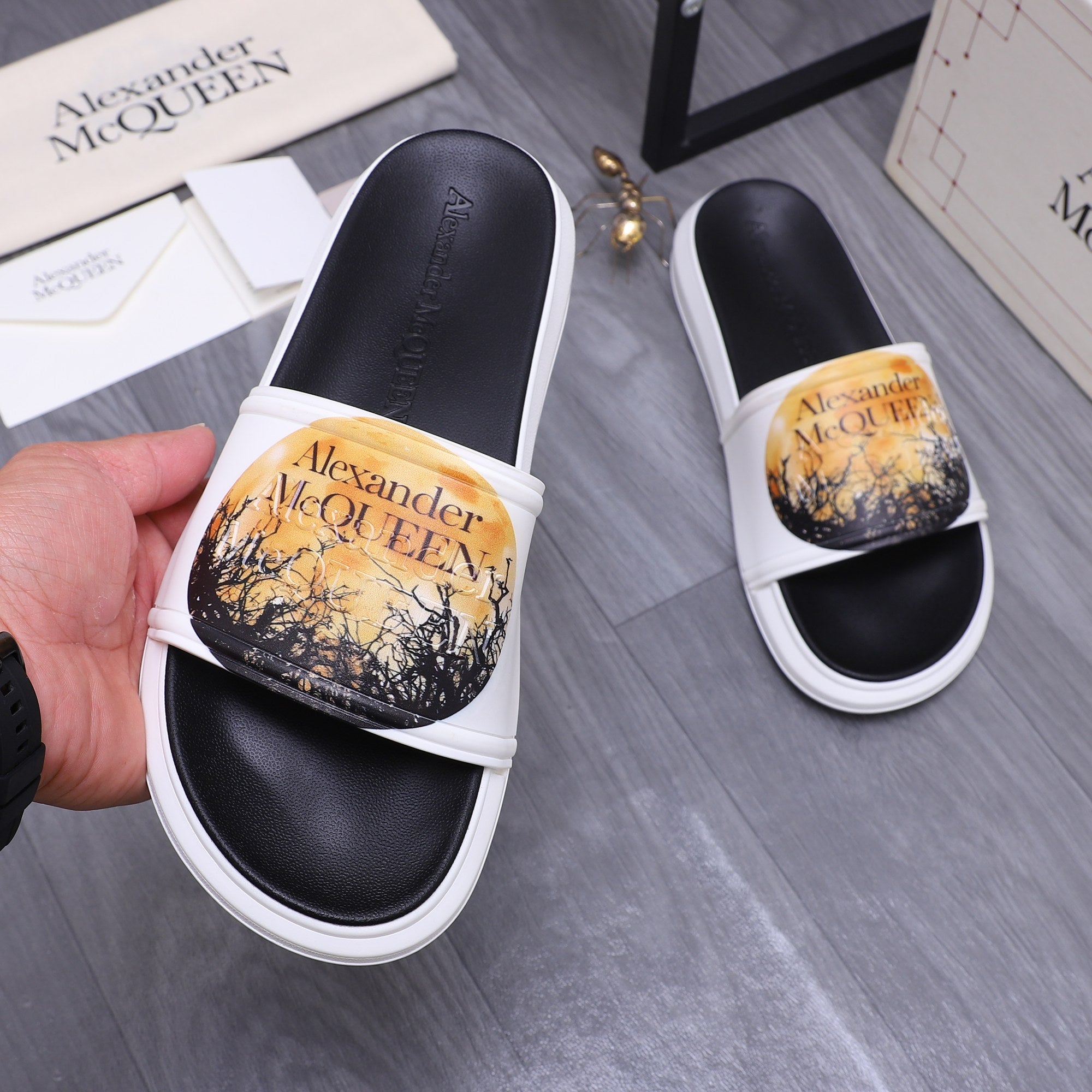 Alexander McQueen slide sandals, black footbeds, white straps, gold graphic print.