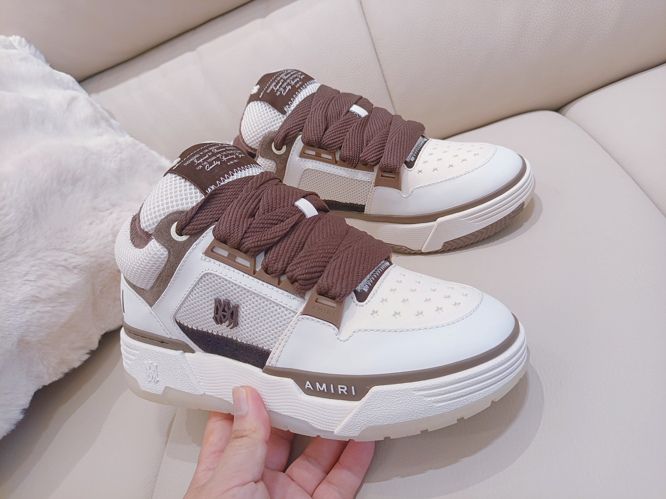 White low-top sneakers with brown laces, held by hand near the front
