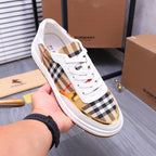 Hand holding a white Burberry sneaker with check upper, gold trim, and orange sole