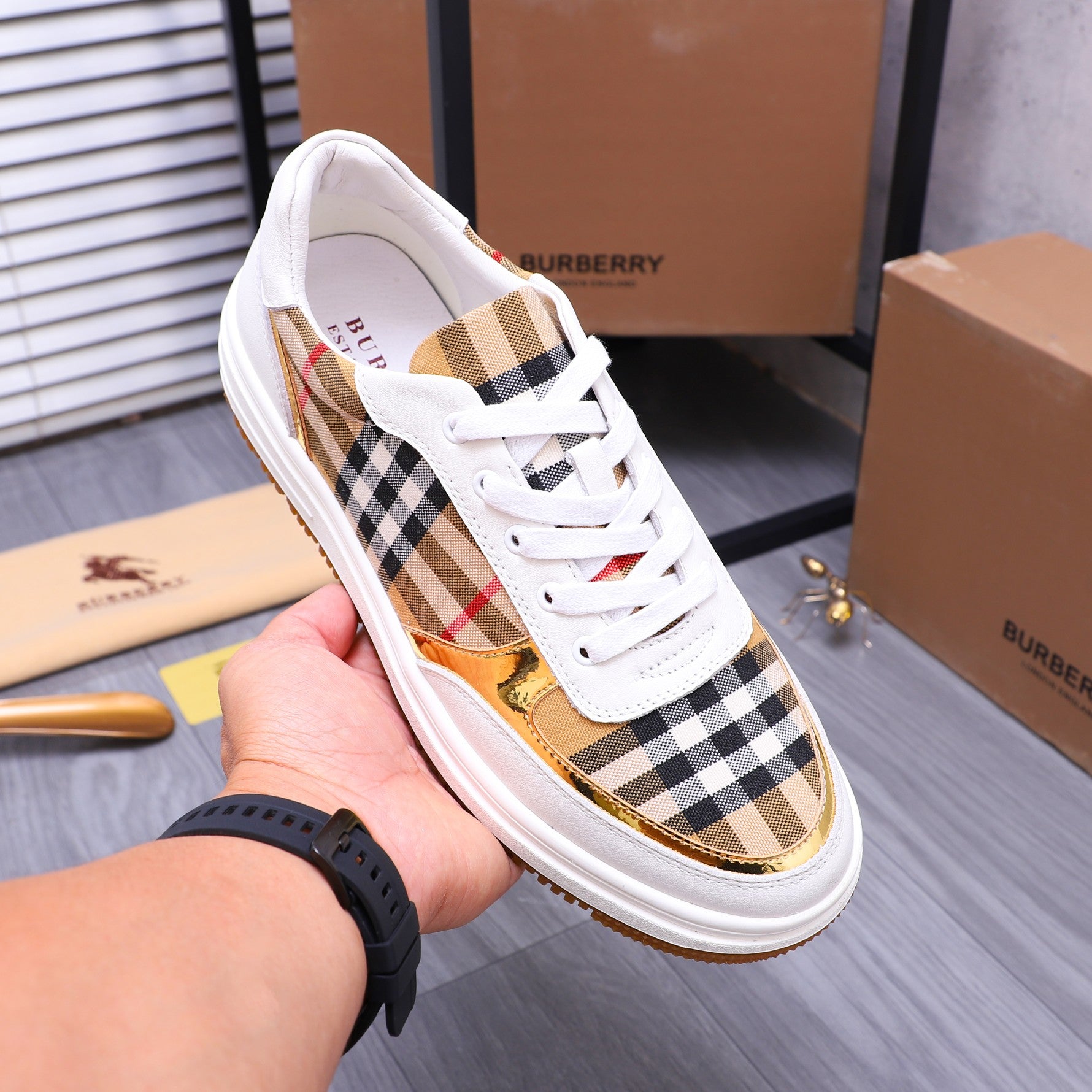 Hand holding a white Burberry sneaker with check upper, gold trim, and orange sole
