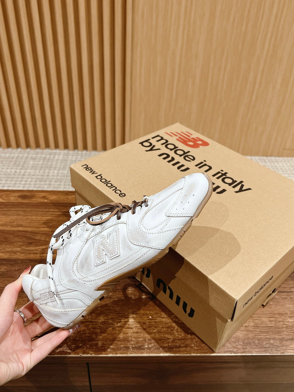 Hand holding white low-top sneakers with brown laces above shoebox
