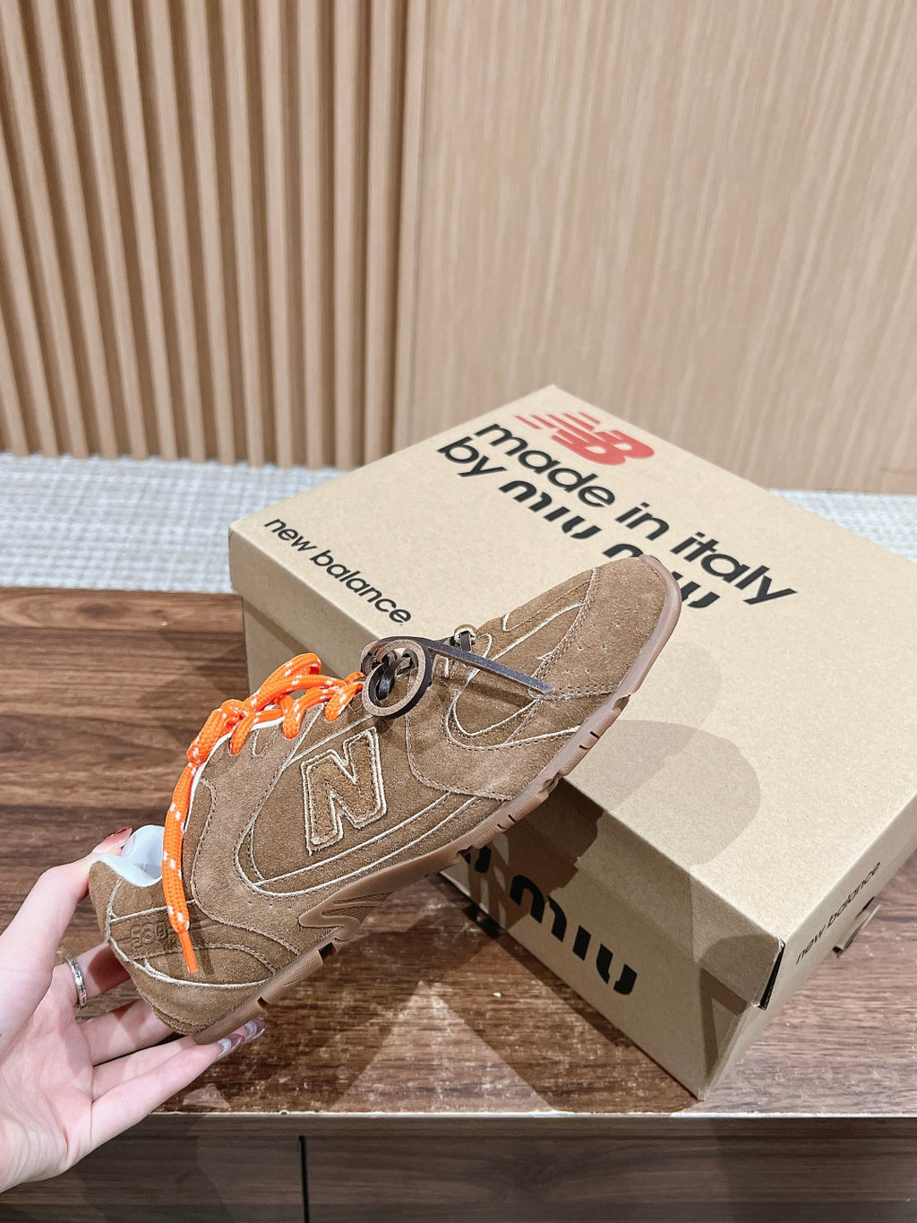 Brown suede sneakers on beige box, orange laces and large N logo on side.