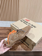 Brown suede sneakers on beige box, orange laces and large N logo on side.