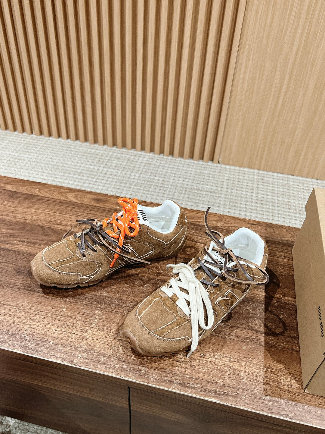 Brown suede sneakers with white laces and orange laces, side view, logo detail.