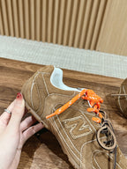 Brown suede sneaker with orange laces held in hand, side profile with N logo.