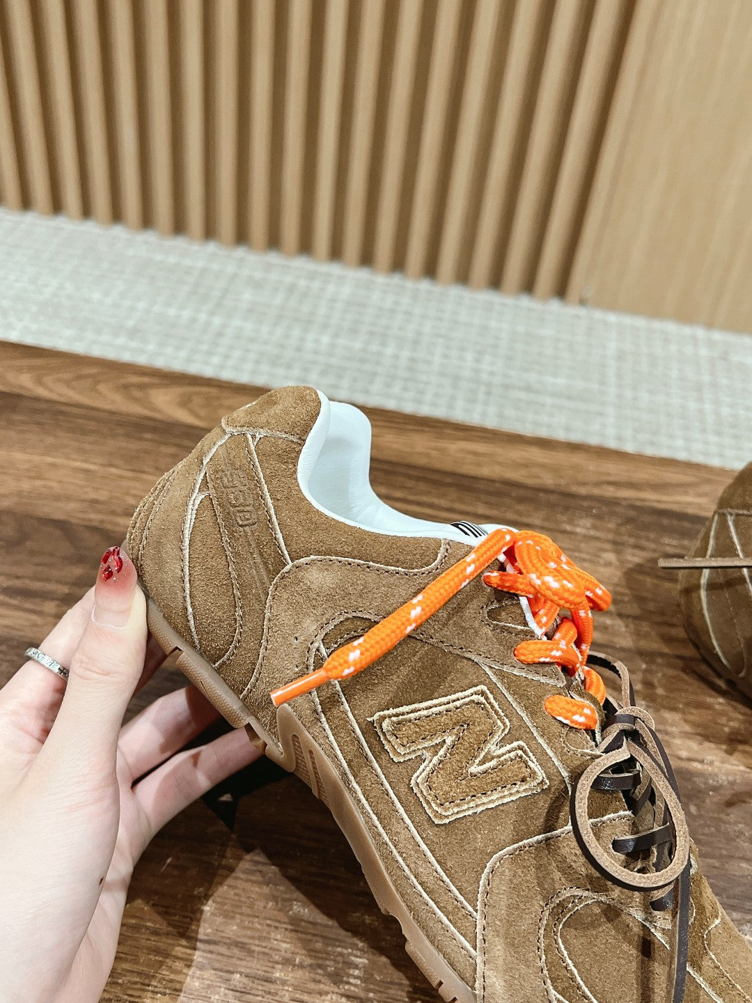 Brown suede sneaker with orange laces held in hand, side profile with N logo.