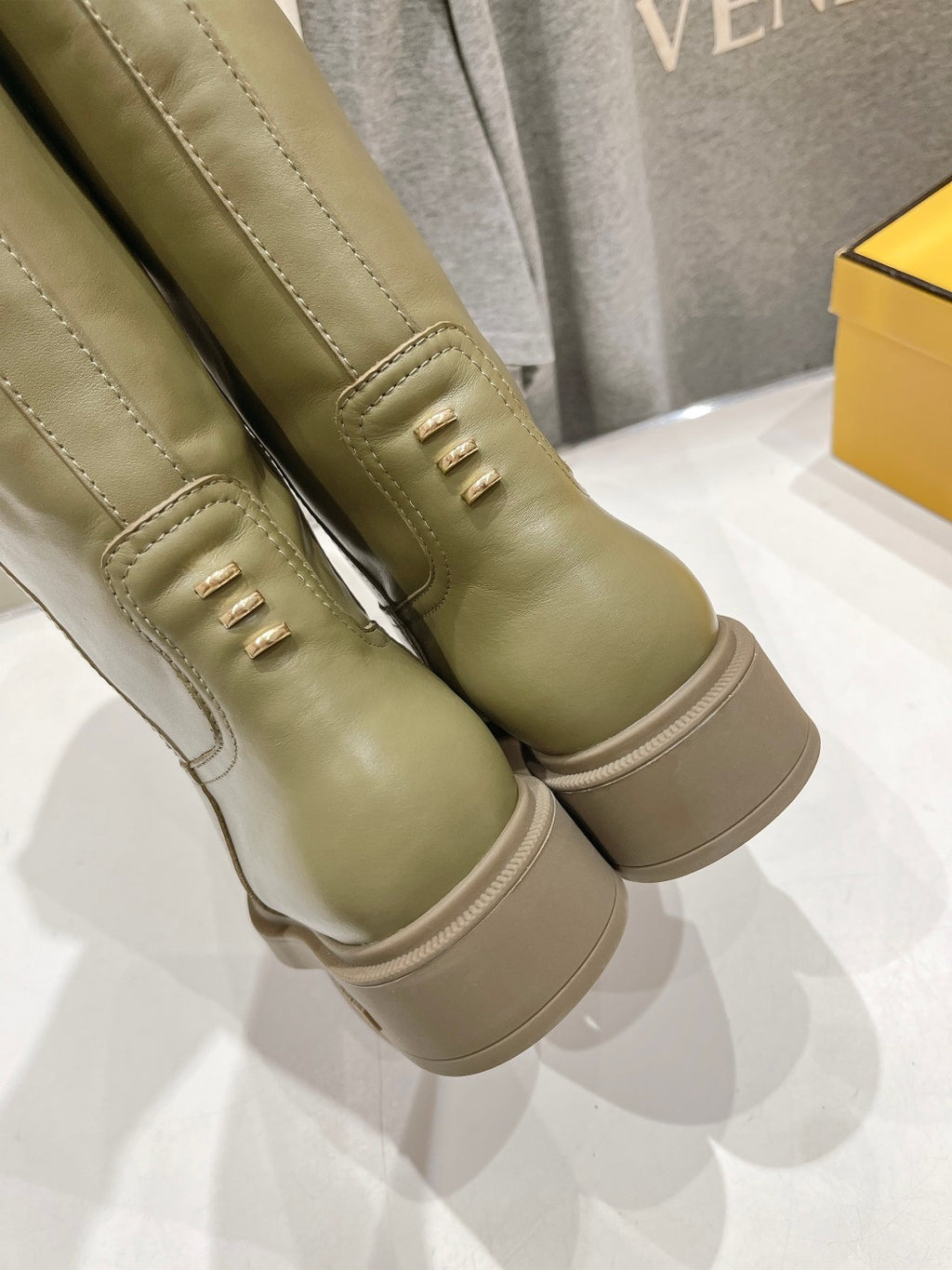 Olive green knee-high boots close-up with stitched panels and decorative eyelets.