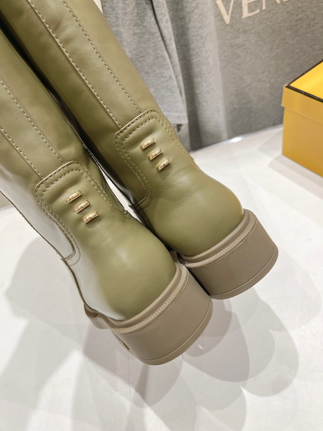Olive green knee-high boots close-up with stitched panels and decorative eyelets.