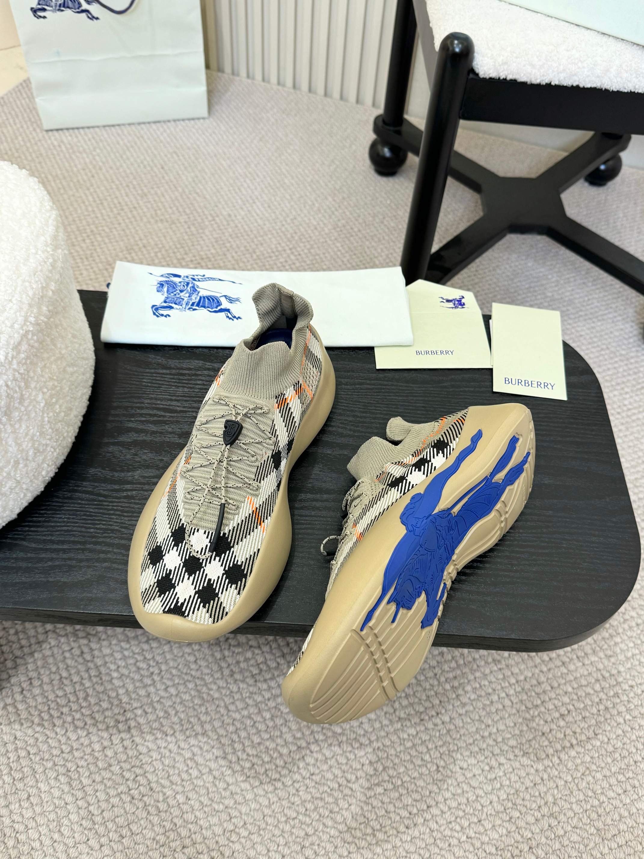 Pair of beige knit sneakers with black check pattern and blue outsoles on table.