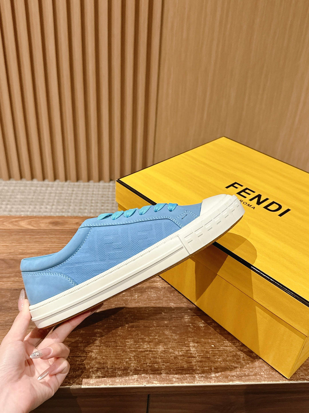Light blue low-top sneakers held in hand with FENDI text on the yellow box.