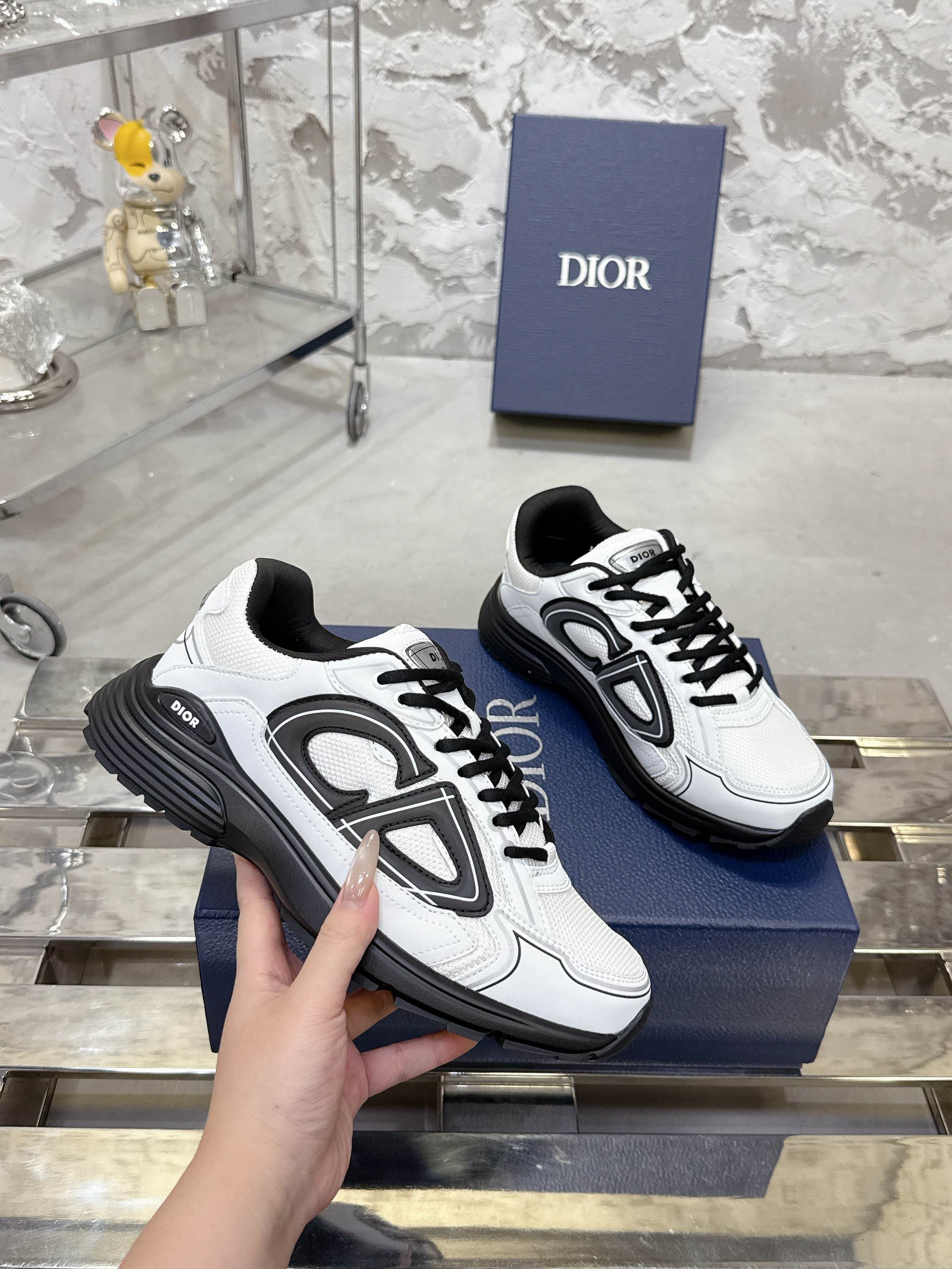 White Dior lace-up sneakers with black chunky soles, hand holding pair on box