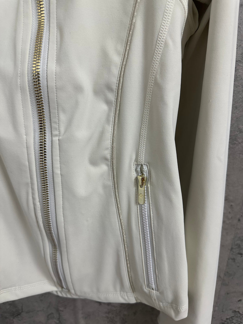 Light white zip-up jacket detail: front zipper with gold hardware and stitched vertical panels.