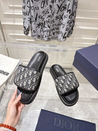 Dior slide sandals being held; black leather-look trim with gray Dior-oblique print