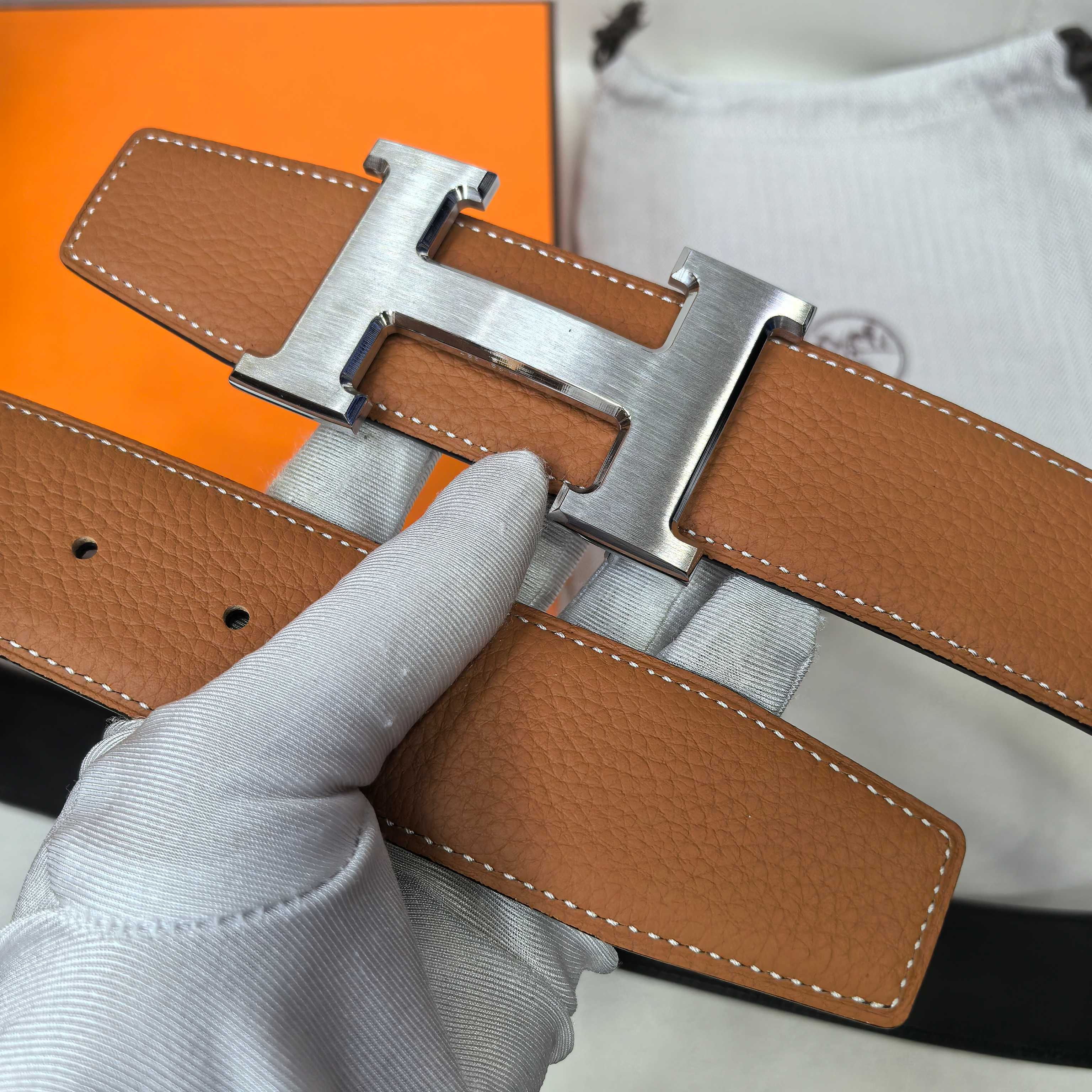 Hermes leather belt close-up with silver-tone buckle, tan-brown strap and holes