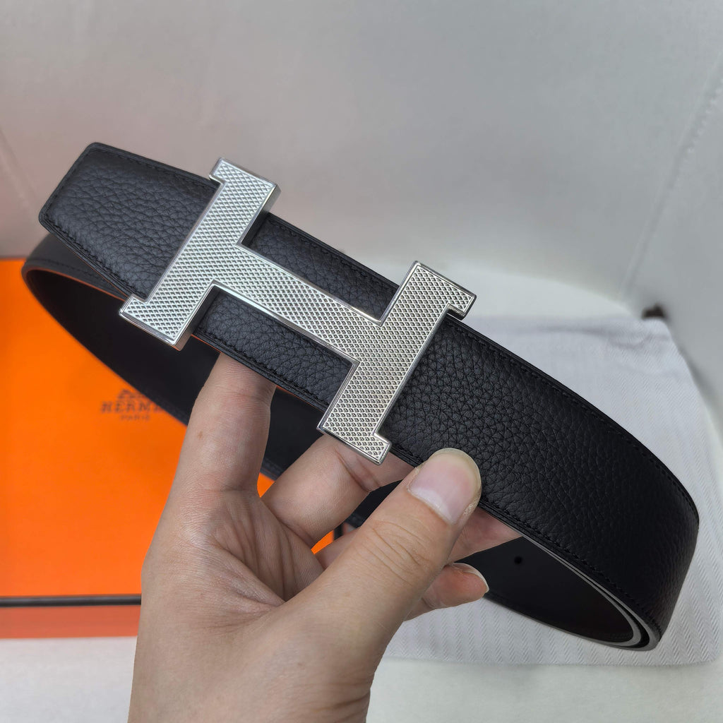 Leather belt with silver H buckle, dark strap with perforation holes, held in hand