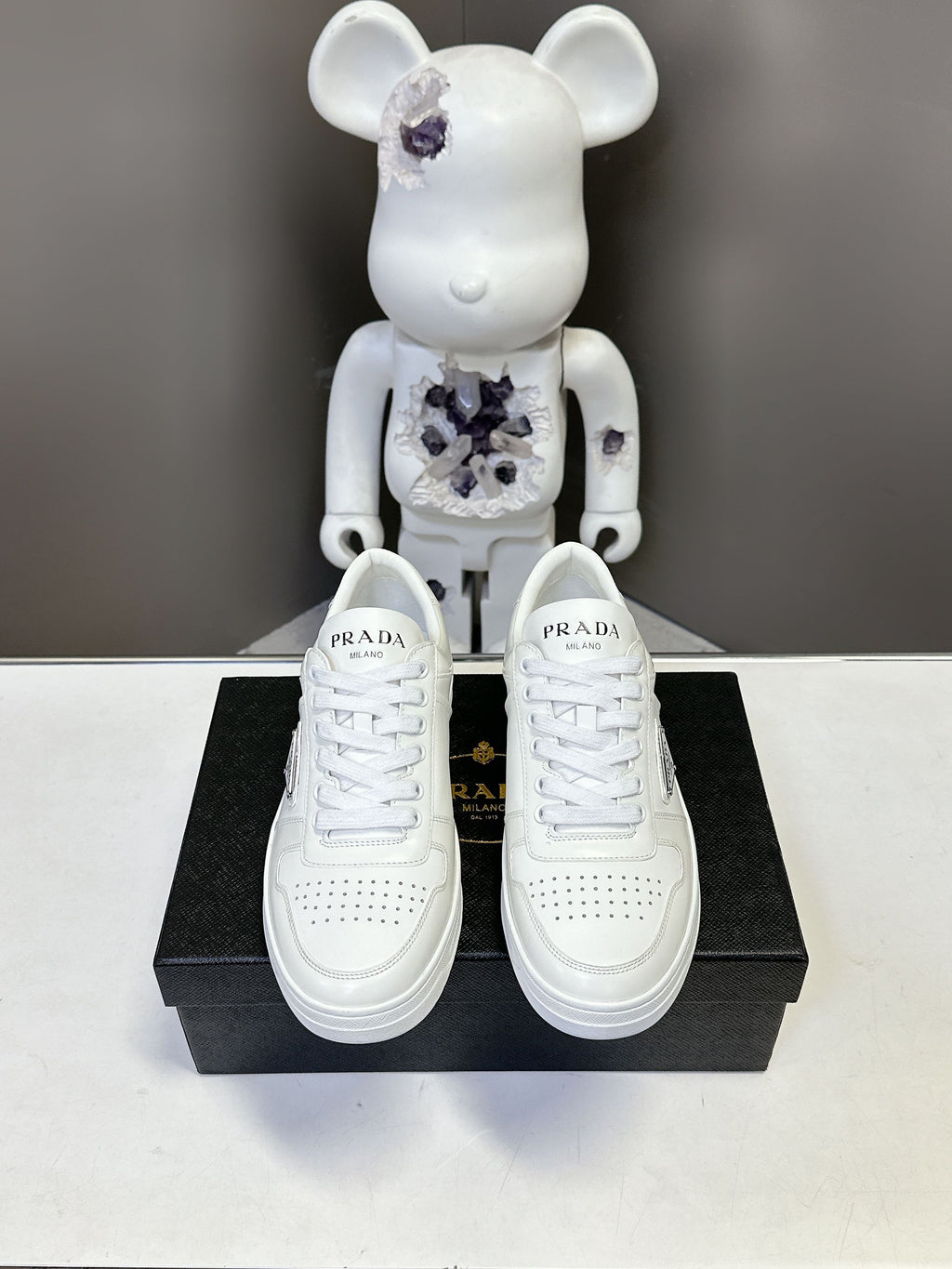 White leather lace-up Prada sneakers displayed on a black shoebox.