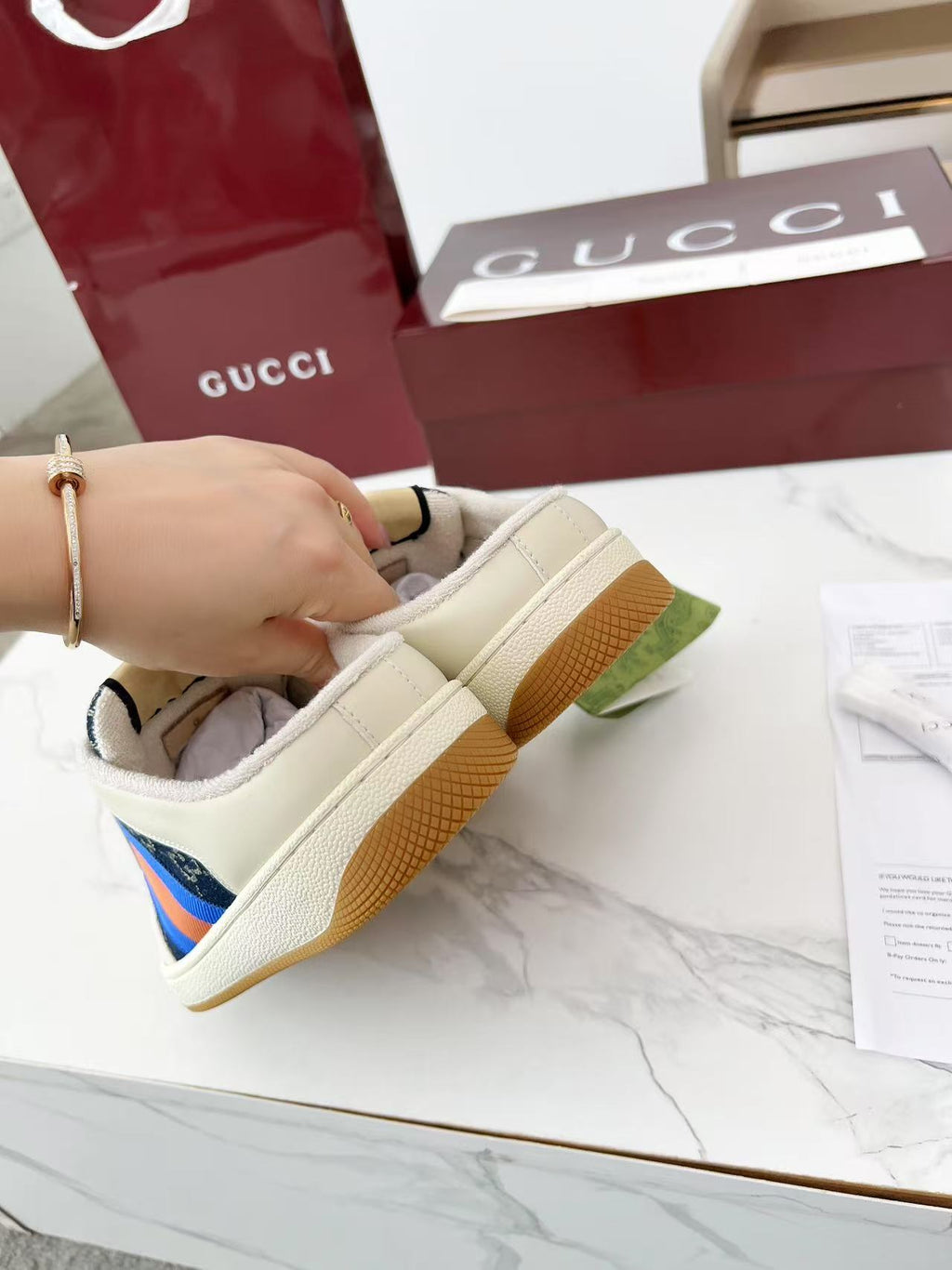 Gucci sneakers held by heel, cream upper with navy GG trim and multicolor stripe