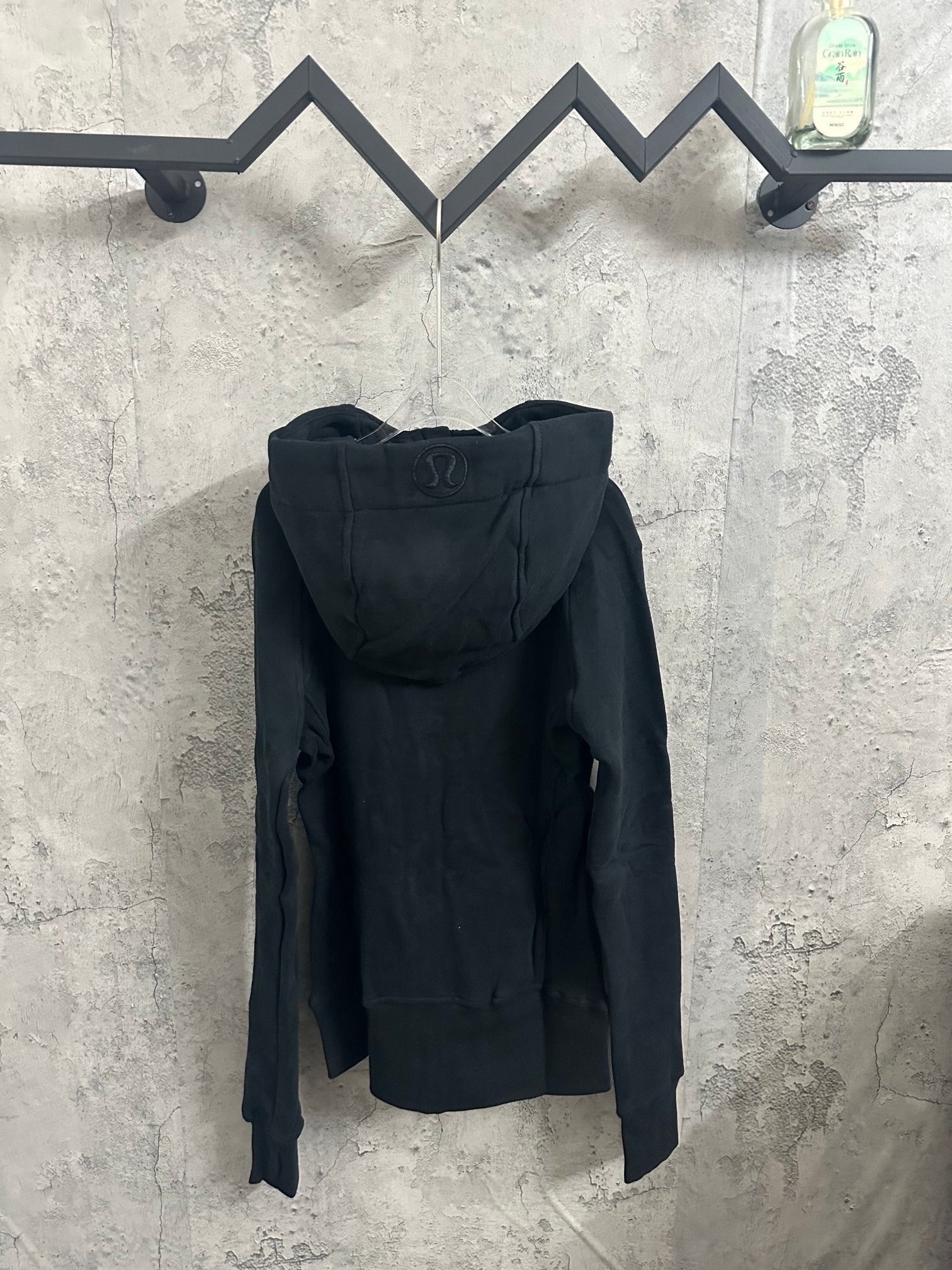 Dark zip-up hooded jacket, back view with hood and drawstrings, hanging retail tag