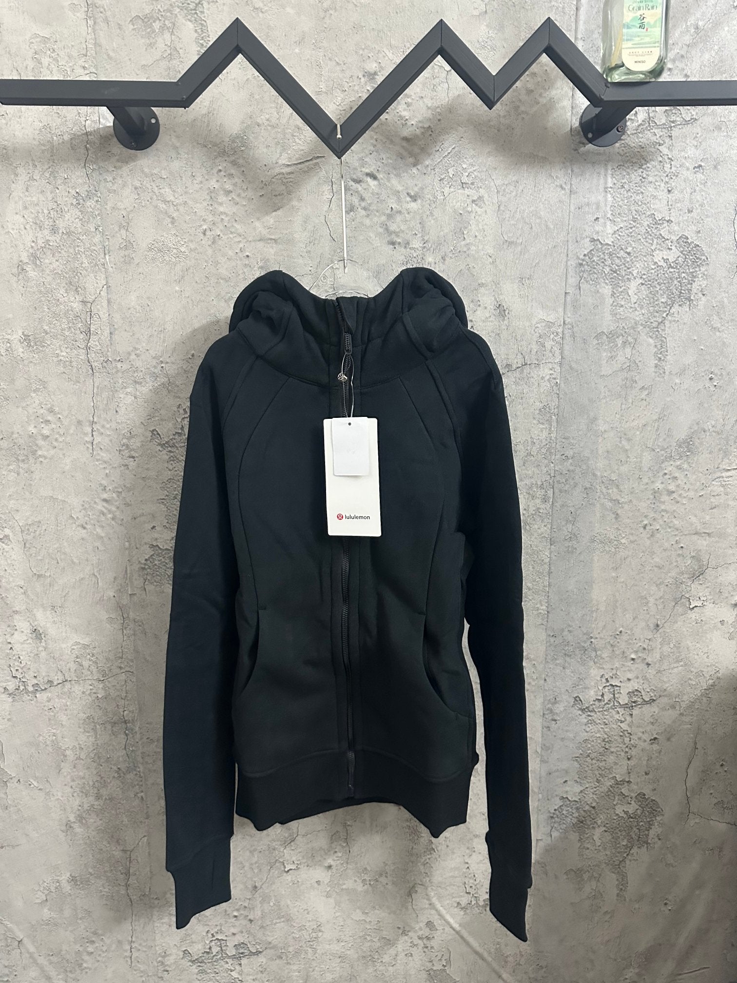 Dark zip-up hooded jacket with front zipper and drawstring hood, hanging retail tag