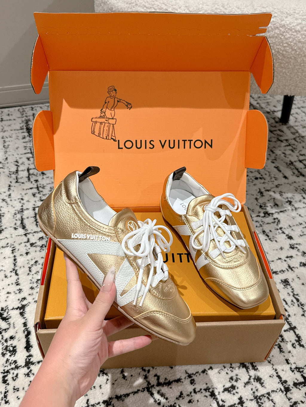 Gold and white leather sneakers with white laces, in open orange shoebox, hand holding.