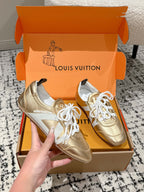 Gold and white leather sneakers with white laces, in open orange shoebox, hand holding.