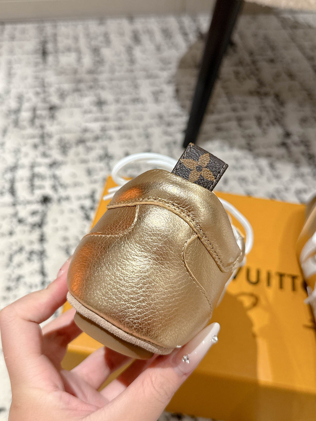 Gold leather sneaker heel with black trim and white laces, held above shoebox.