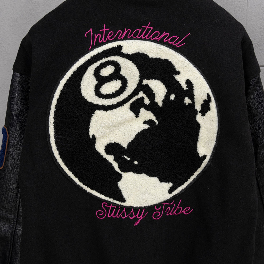 Black varsity jacket back with large globe patch and pink “Stussy Tribe” text