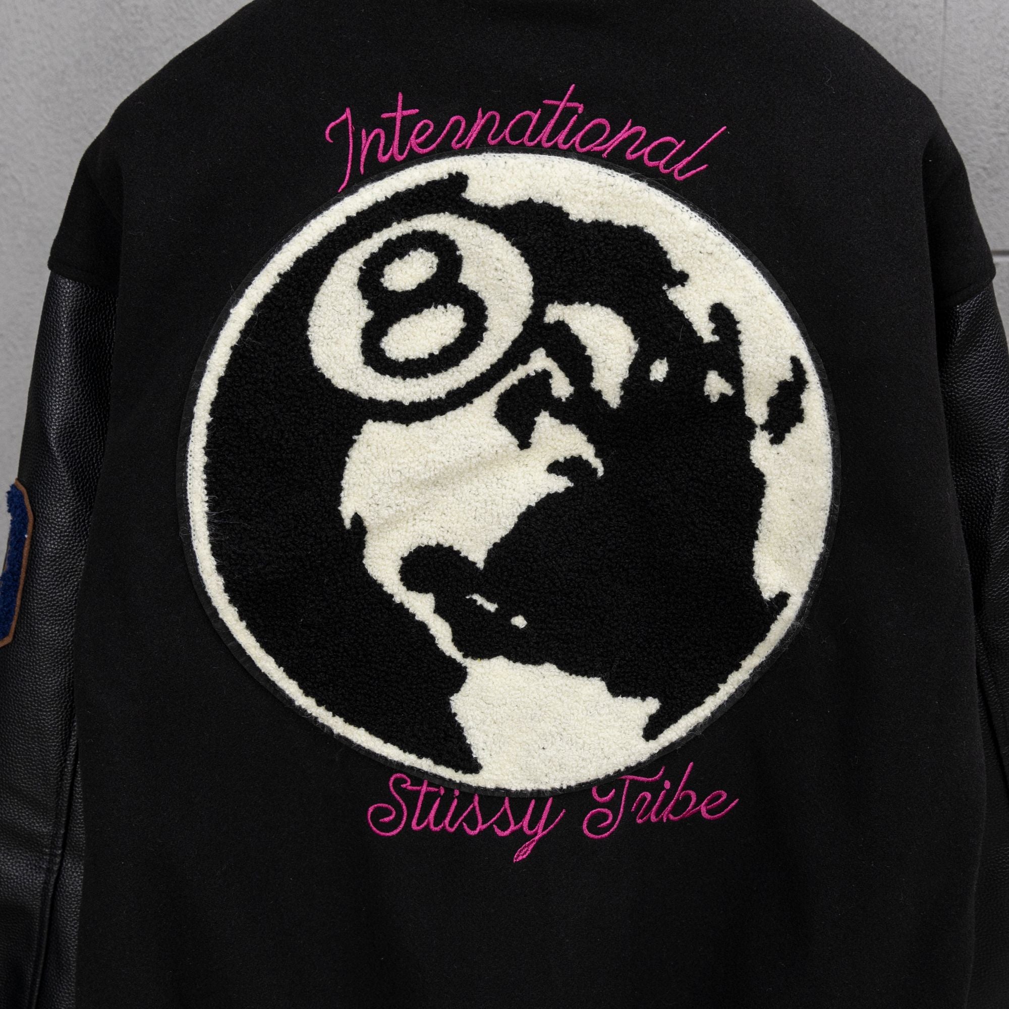 Black varsity jacket back with large globe patch and pink “Stussy Tribe” text