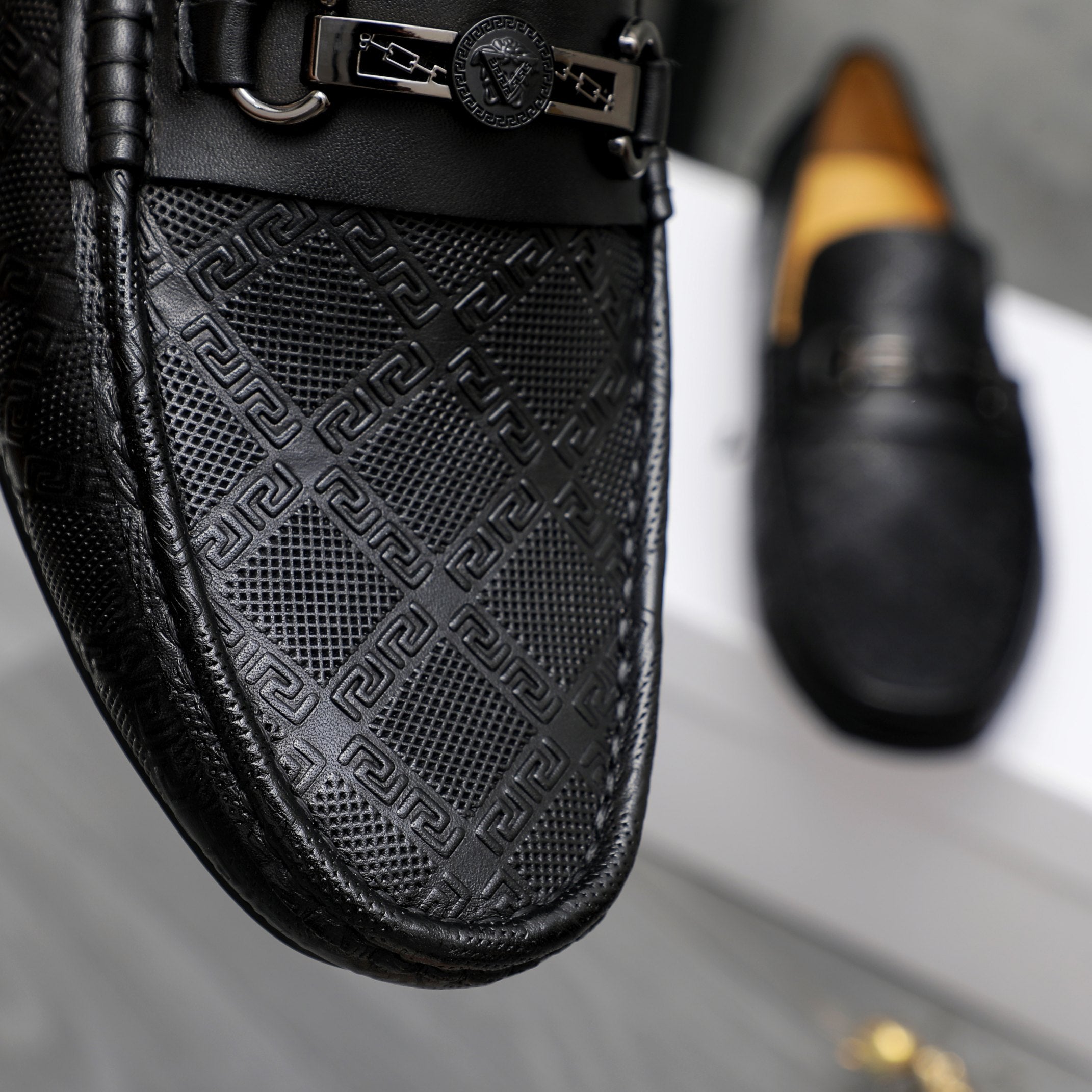 Close-up of black loafer upper with embossed geometric pattern and metal buckle strap.
