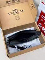 Small black crossbody bag with zip top and front pocket, shown in a box