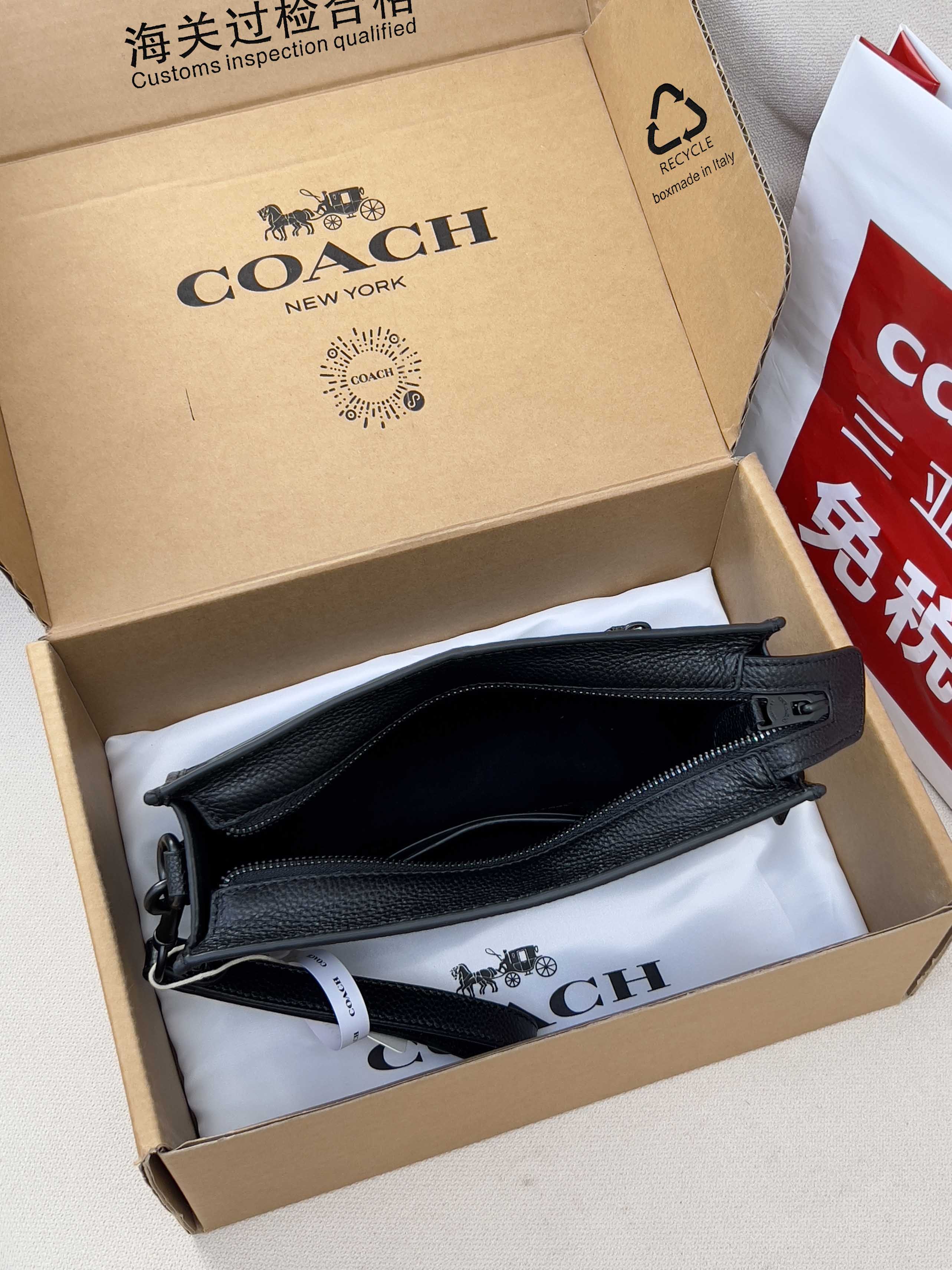 Small black crossbody bag with zip top and front pocket, shown in a box