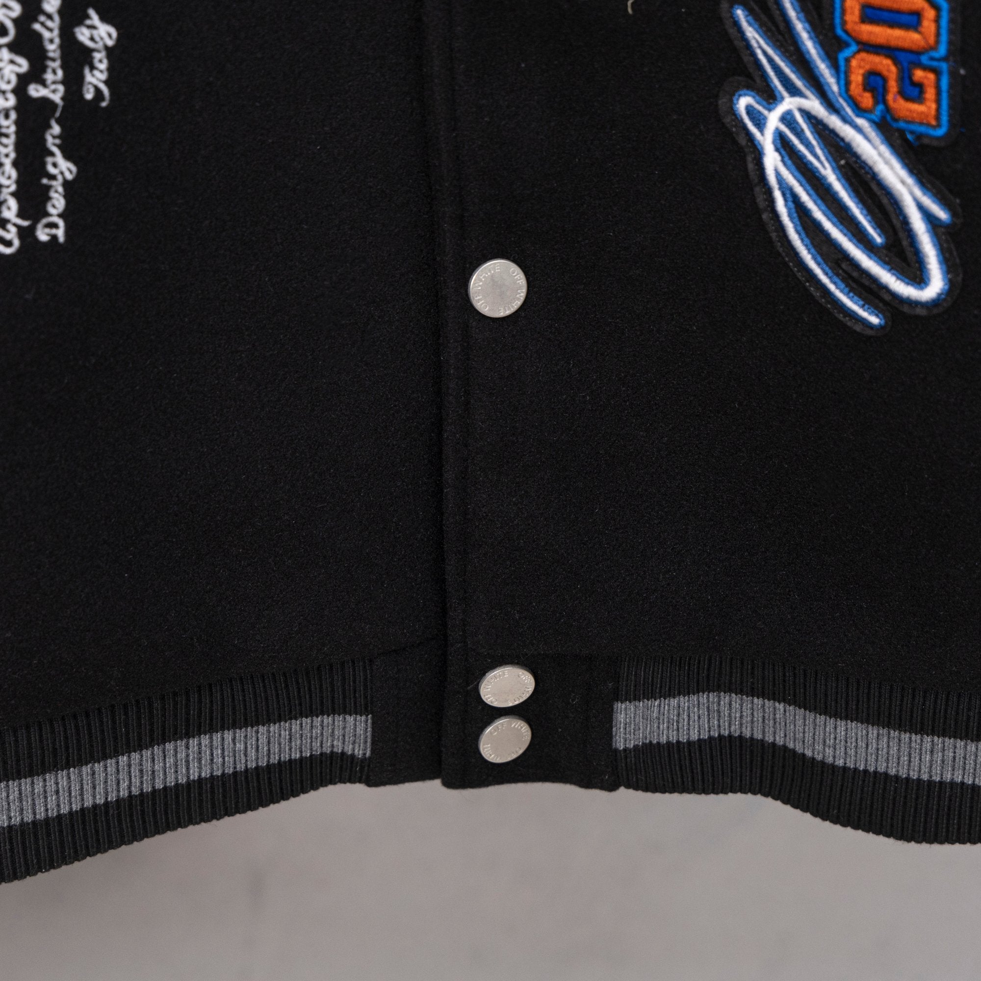 Black varsity jacket front with snap buttons and striped ribbed hem trim.