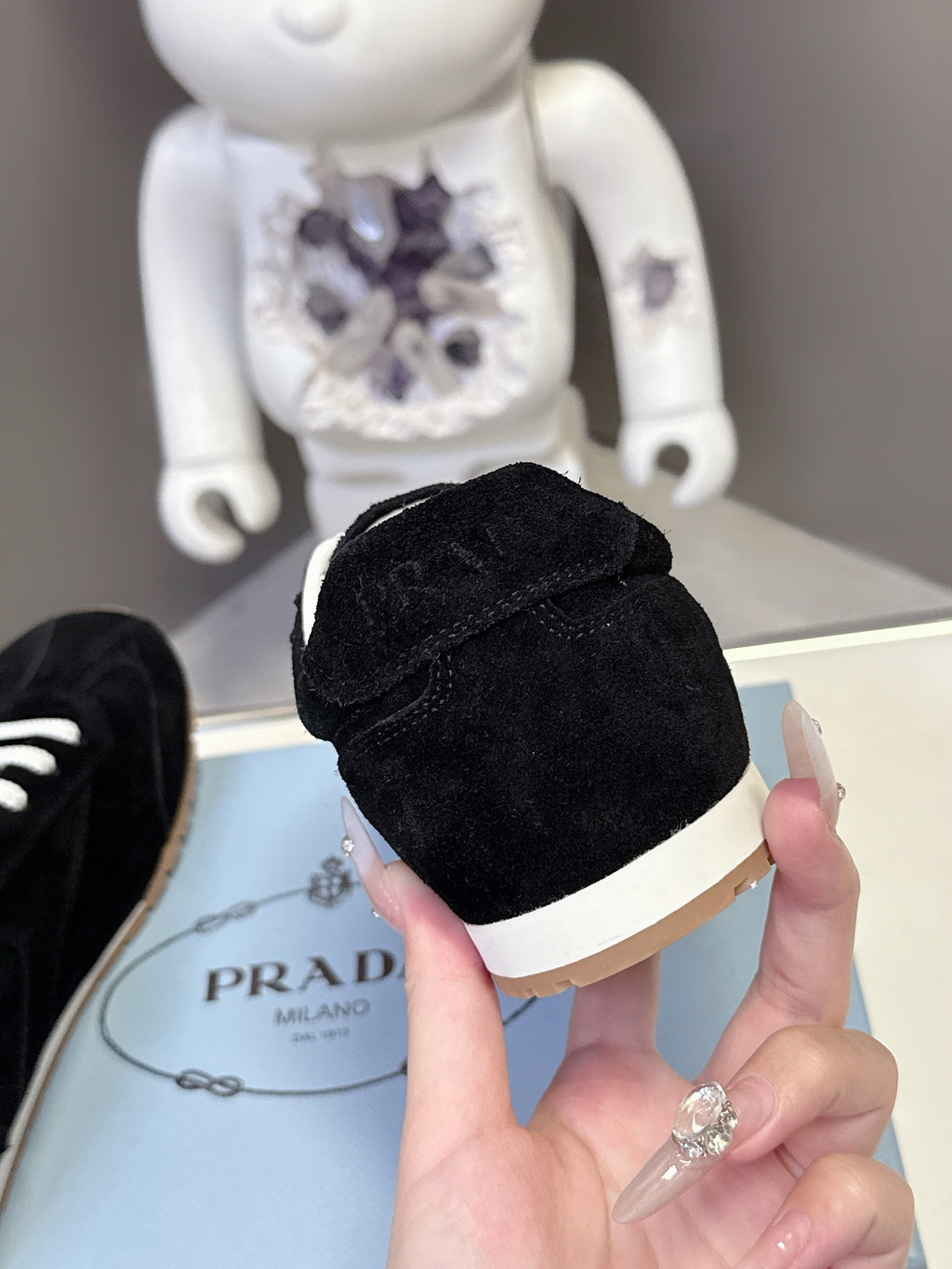 Black suede sneaker toe with white sole edge, held above Prada shoebox.