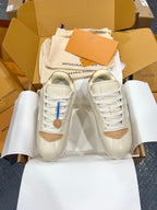 Cream low-top sneakers in a box with white laces, tan trim, and logo on the tongue