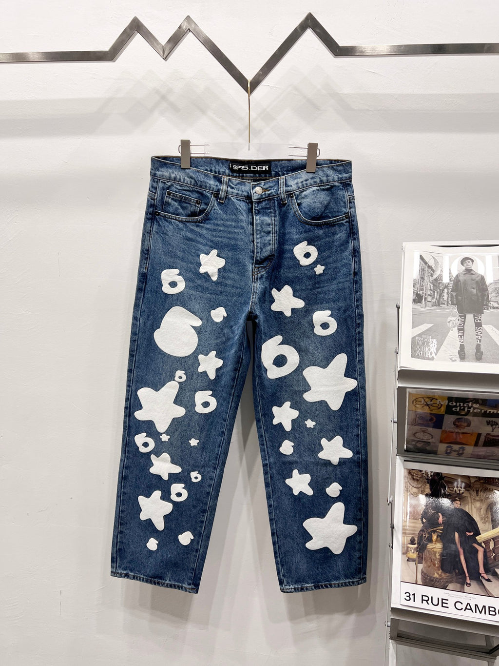 Blue denim pants with white graphic patches, front view on hangers