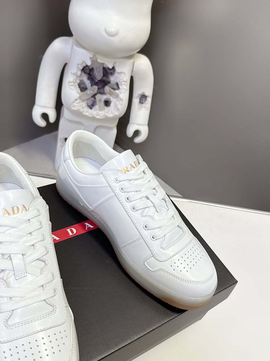 White Prada sneaker side view with laces and stitched overlays, on a black box