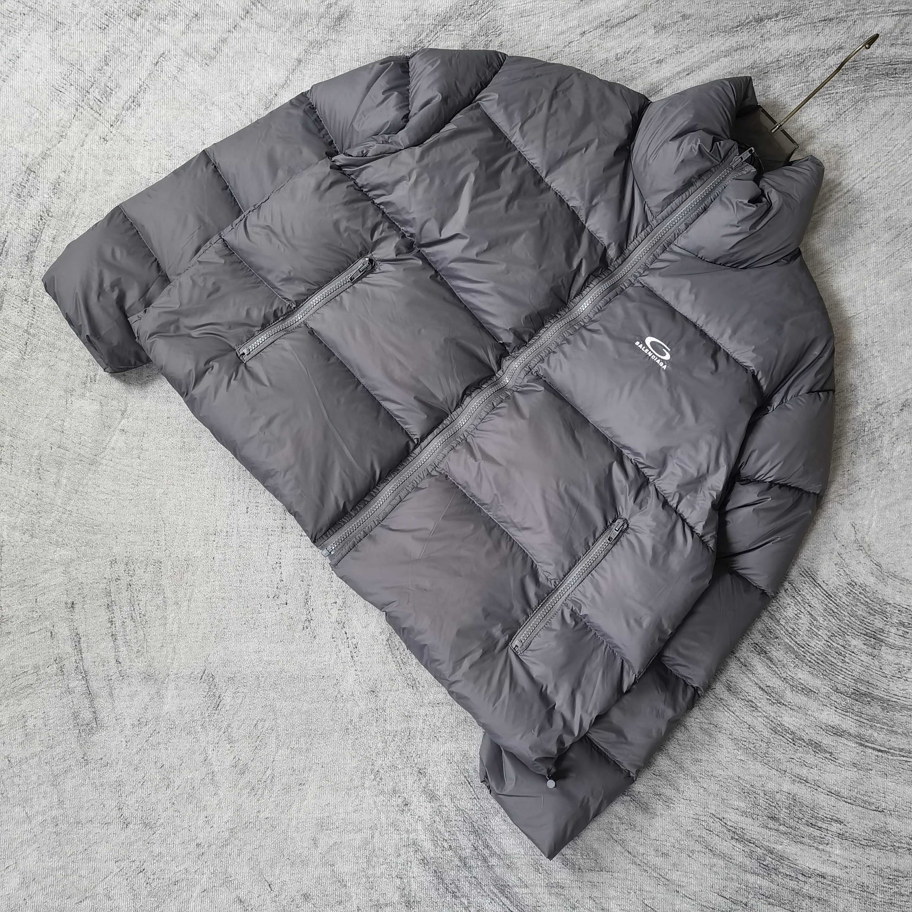 Gray Balenciaga puffer jacket laid out, angled view with zip front and chest logo.