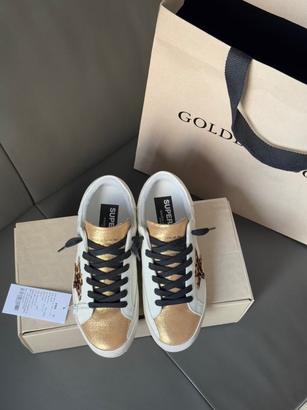 Two white sneakers with black laces and gold toe caps on a shoebox