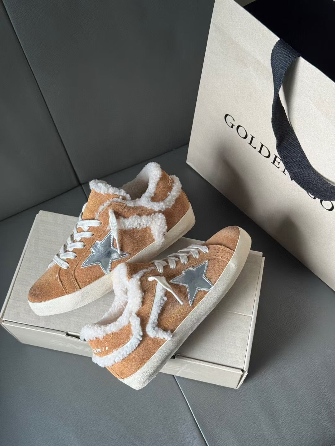 Brown suede sneakers with white shearling trim and blue star patches, shown on a box.