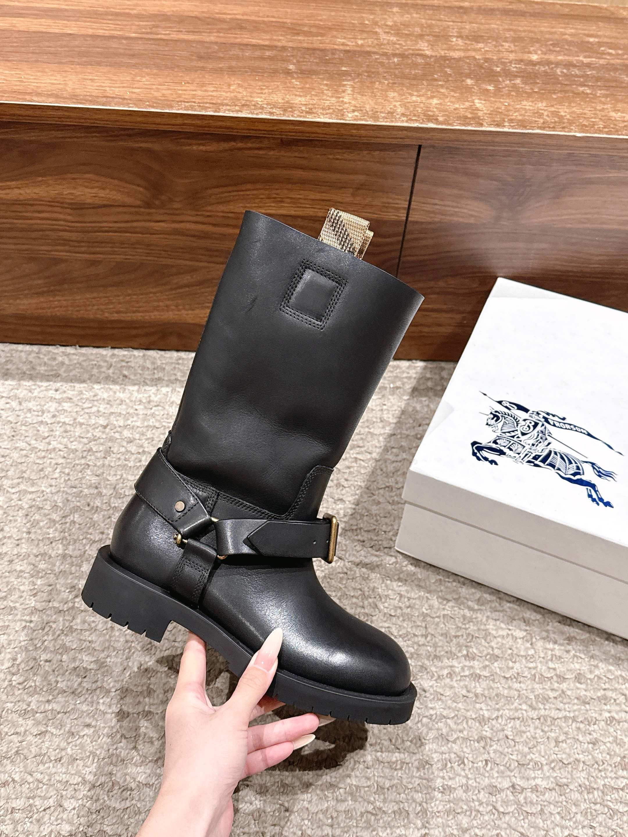 Black mid-calf boot held in hand, with front strap buckle and chunky sole.