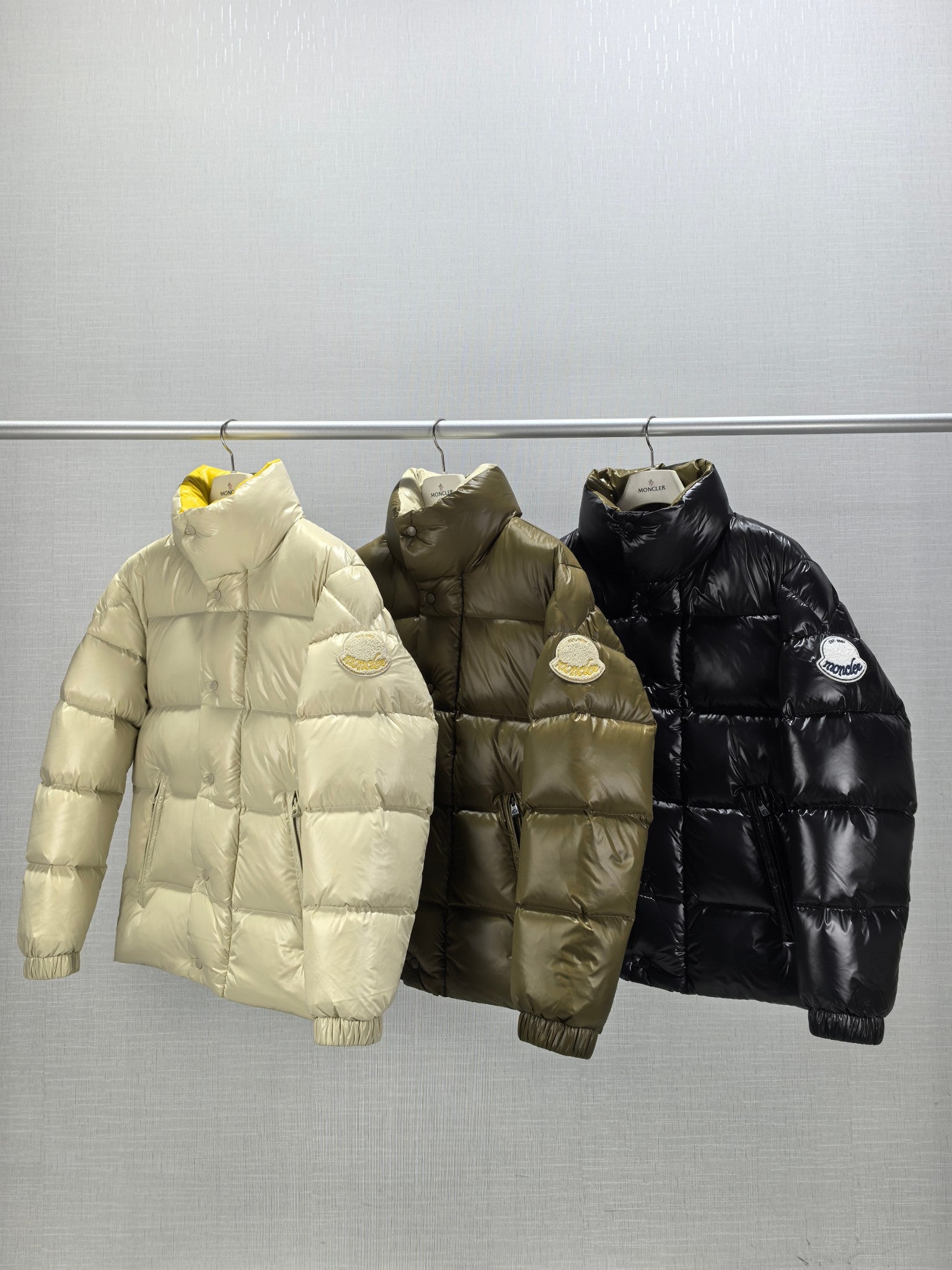Three Moncler puffer jackets: light, olive, and black on hangers