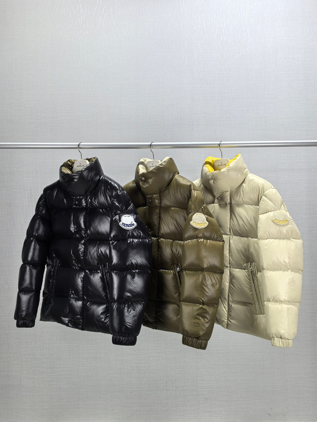 Three Moncler puffer jackets on hangers: black, olive, and light, front view
